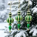 Set di tre ornamenti natalizi a strisce verdi e oro appesi a un ramo di pino innevato - Verde