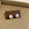 Close-up of elegant white pearl stud earrings with a lustrous finish on a wooden base. - Imitation Shell Pearl (HE8071) / 14mm Size