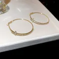 Detalle de primer plano de elegantes pendientes de aro dorados con cristales pavé y motivo en T - Dorado