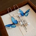 Close-up of elegant blue butterfly earrings with white pearls resting on a wooden display tray - Ear Threader Style (HE20215-HE20216) / Silver-Blue
