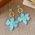 Close-up detail of blue resin flower earrings with textured gold hoops on a woven background - Resin Flower Pendant Style / Gold Tone And Blue