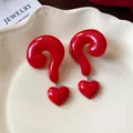 Detail of glossy red question mark earrings with heart charms on a ceramic plate. - Ancora Red (Silver Pin)
