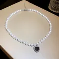 Elegant close-up of a white pearl necklace featuring a black heart-shaped pendant detail. - Black (Heart)