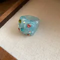 Close-up of a light blue resin ring with colorful rhinestones on a textured display. - Cyan