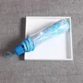 Compact light blue folding umbrella featuring daisy patterns in a clear plastic pouch - Manual Sakura Blue Transparent