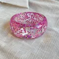 Close-up of a chunky transparent resin bangle with embedded pink iridescent glitter. - Purple