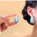 Dettaglio di orecchini a bottone in resina azzurra a forma di C su orecchio e mano - Blu