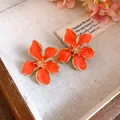 Close-up of elegant orange enamel flower earrings with gold trim and textured centers. - Tops / Orange