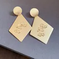 Macro view of gold rhomboid pendant earrings featuring an elegant embossed floral pattern - Silver Earring Post Style / Gold