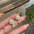 Hand showing elegant white flower stud earrings with shimmering zircon crystal drop - Flower Shaped Style (HE34453) / Silver (White Petals)