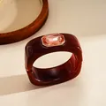 Dettaglio di un bracciale quadrato in resina rosso scuro con una gemma color pesca lucida. - Marrone