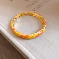 Primo piano di un bracciale smaltato arancione e giallo su sfondo di un libro beige - Oro, Giallo e Arancione