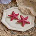 Detail view of pink starfish stud earrings with gold trim and pearl centers on a straw mat - Rose Red
