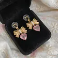 Close-up of golden bow earrings with pink heart crystals and pearls in a black velvet box - Ear Clip - Gold - Pink (Real Gold Plated)