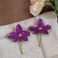Elegant purple enamel orchid earrings with rhinestones on a beige textured pouch background. - Purple
