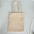 Plain beige canvas tote bag laid flat on a textured white fabric background detail - Apricot (8oz)