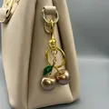 Nahaufnahme eines Kirsch-Taschenanhängers an einer beigen Lederhandtasche. - Gold mit Grünem Blatt