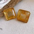 Dettaglio macro di orecchini a bottone quadrati in acrilico ambra con cornice dorata - Stile con perno dell'orecchino in argento / Marrone caffè