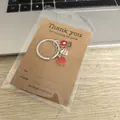 Gros plan détaillé d'un porte-clés pomme et fleur sur un support cartonné brun - Grande Carte + Sac OPP / Rouge