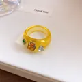 Detailopname van een modieuze gele doorschijnende harsring met kleurrijke strassteentjes. - Zetting met drie diamanten / Geel-Koffie