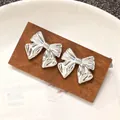 Elegant silver metallic bow-tie stud earrings displayed on a dark wood block. - 3D Pleated Bowknot Stud Earring (N-355)