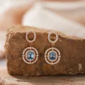 Nahaufnahme eleganter goldener Ohrhänger mit ovalem Blautopas im Halo-Design - Natürlicher Topas Ohrstecker / Gold