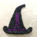 Nahaufnahme eines funkelnden schwarzen Hexenhutes aus Lametta mit lila BOO-Schriftzug. - Hexenhut