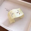 Macro detail of cream acrylic cuff bracelet featuring elegant hollow floral cutout patterns. - Yellow