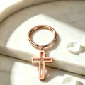 Primer plano elegante de un llavero con cruz hueca de oro rosa sobre mármol blanco - Rosa oro