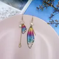 Nahaufnahme asymmetrischer bunter Zikadenflügel-Ohrringe mit goldener Herzquaste - Regenbogen Strass-Gold