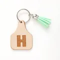 Detail of a handmade leather ear tag keychain featuring letter H and a mint green tassel - Letter H Style (20438)