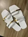 Close-up of white flat sandals with elegant gold metal buckle straps on wood floor. - White