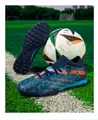 Close-up detail of blue flyknit soccer cleats with TF studs on a green grass field background - Navy Blue & Orange Turf Cleats