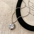 Détail d'un collier pendentif montre en forme de cœur argenté sur fond beige texturé. - Cœur / Argent