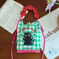 Close-up of a cartoon cat mini crossbody bag with green gingham and a red handle accent. - Black Cat
