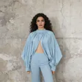 Model draagt een lichtblauwe cape blouse met pofmouwen en bijpassende broek bij een muur - S / Tops / Lichtblauw