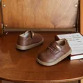 Nahaufnahme von braunen Kinder-Brogue-Lederschuhen auf einem Holzstuhl neben einem Buch - 16 / Braun