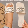 Detail view of cream square toe flat sandals featuring a sparkling rhinestone buckle. - Beige