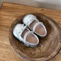 Zapatos Mary Jane plateados para niña con perlas y lazo, presentados en un cuenco de madera. - Plateado