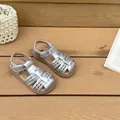 Detalle de sandalias trenzadas plateadas para niña sobre fondo de madera. - Plateado