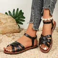 Stylish black flat Roman sandals for women with criss-cross straps on a gravel beach. - Black