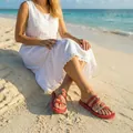 Red and gold flat casual sandals worn by a woman sitting on a sunny sandy beach scene - 38 / Red