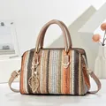 Detalle de bolso tipo almohada tejido a rayas con dije de hoja dorada y asa superior. - Caqui