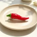 Detailed view of a realistic red chili pepper hair clip on a minimalist beige plate. - Chili