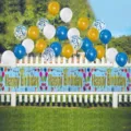 Blue Happy Birthday fringe banner with assorted balloons displayed on a white fence - Text Style / 30x270cm / Sky Blue