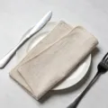 Beige overlocked cotton linen cloth napkin on a white dinner plate with silver cutlery - 32x32cm / Beige
