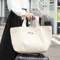 Bolso tote de lona blanca grande con texto 'HER' sostenido por una mujer en viaje - Estilo Tote con Asa y Letra HER (SR6679)