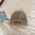 Close-up of a soft beige knitted beanie hat with embroidered text on white textured cloth - One Size / Khaki