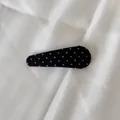 Détail d'une pince à cheveux noire à pois blancs sur un fond en tissu texturé - Barrette Goutte d'Eau / Noir