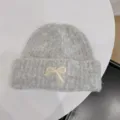 Detail of a grey knitted beanie featuring a cream bow applique on a white table - One Size / Gray